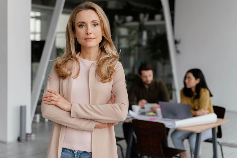 Une chef d'entreprise sereine et souriante, debout devant son équipe travaillant en arrière-plan, illustrant la protection d'une garantie frais professionnels.